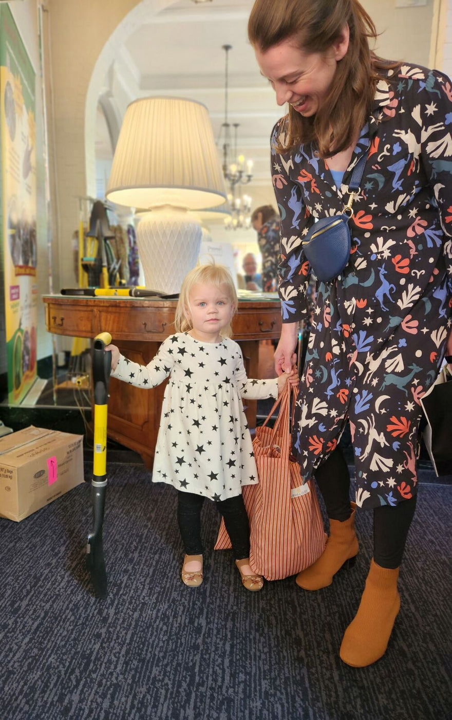A toddler in a star-print dress stands beside a smiling parent, one hand on a yellow Spear Head Spade, at PIZZAZZ! 2026.